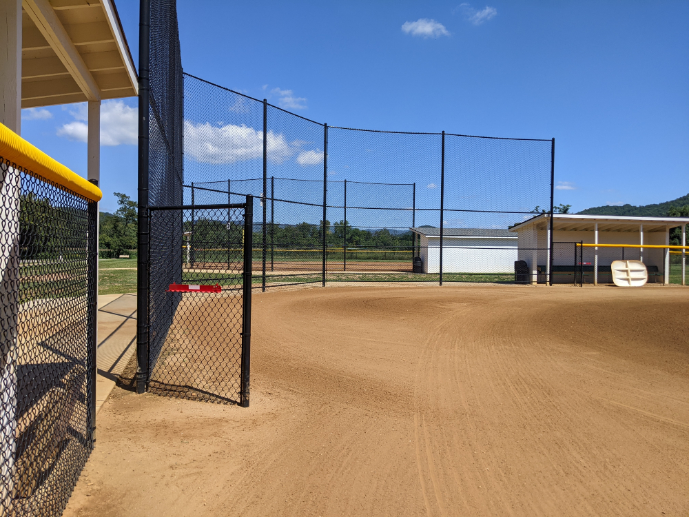 The backstop of one of the ballfields is shown from the first base side on a sunny summer day.