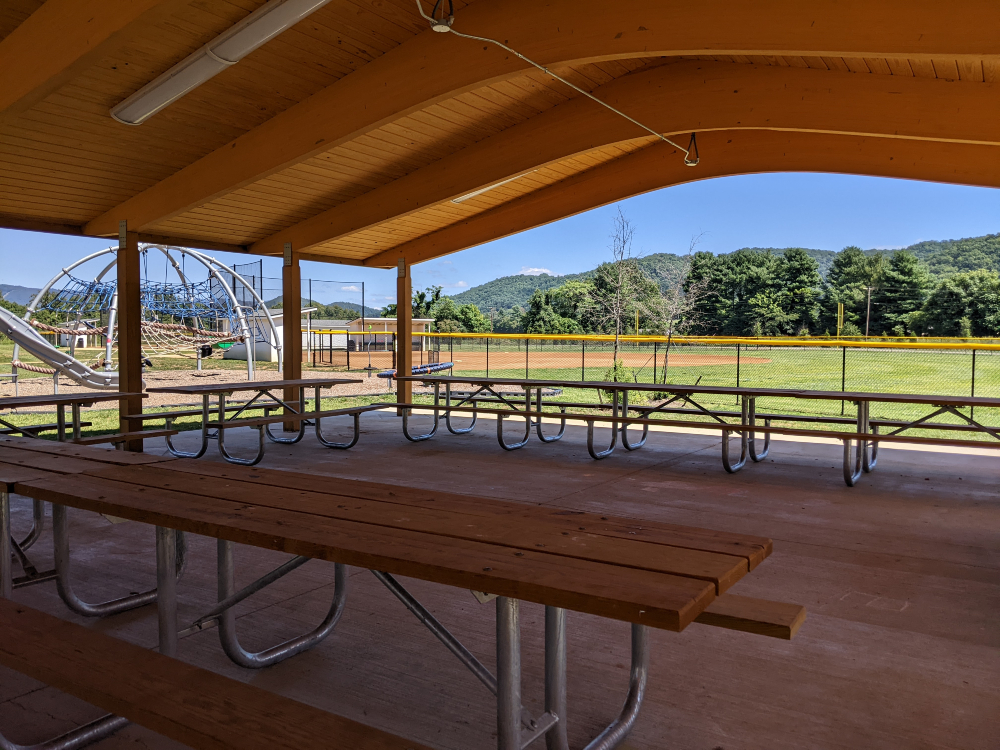 A view from under the picnic shelter shows the playground and ball fields with mountains and blue sky in the distance on a sunny summer day.