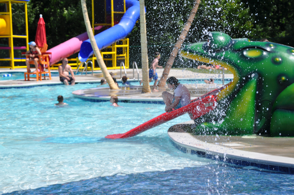 In the foreground, water falls from a spray feature while a child slides down the Frog Slide at the Frog Pond Swimming Pool on a sunny summer day.