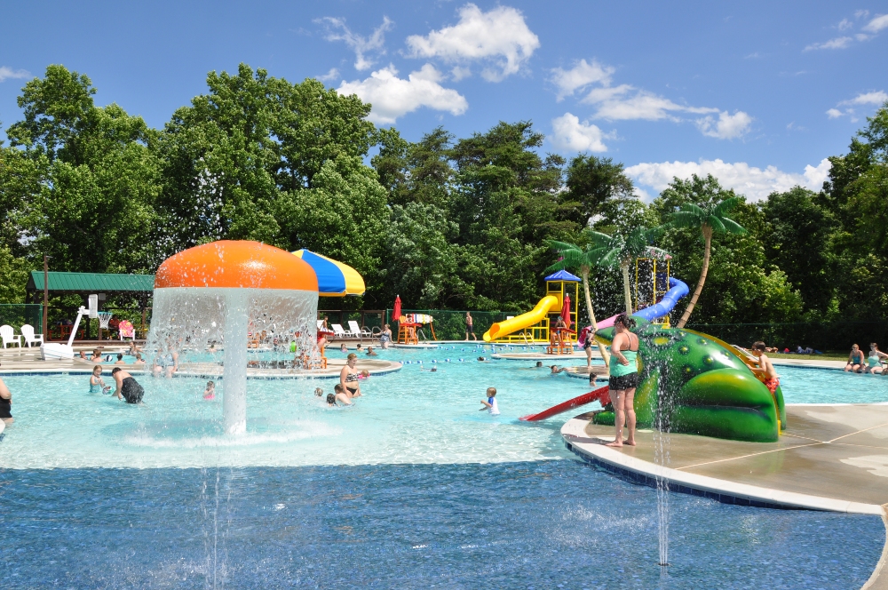 Citizens are seen using splash features, slides, a water basketball goal, and sun bathing areas at the Frog Pond Swimming Pool on a sunny summer day.