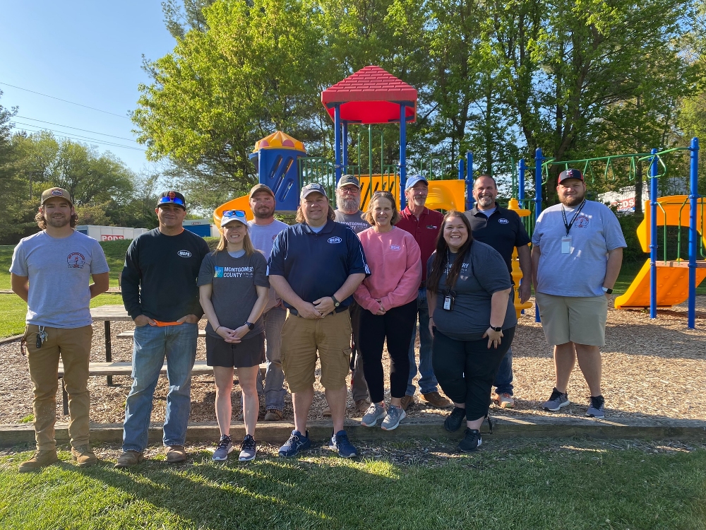 Montgomery County Parks and Recreation staff pose for a group photo in front of a playground and trees on a sunny summer day.