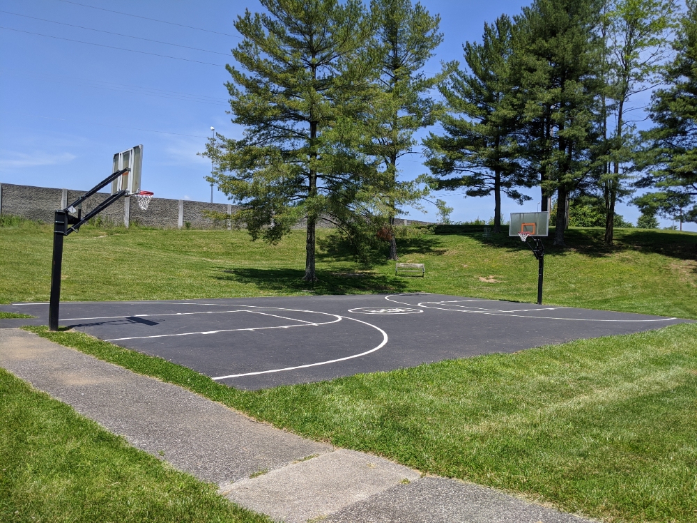 The outdoor basketball court at Mid County Park is surrounded by green space and pine trees, and has a bench to rest on.