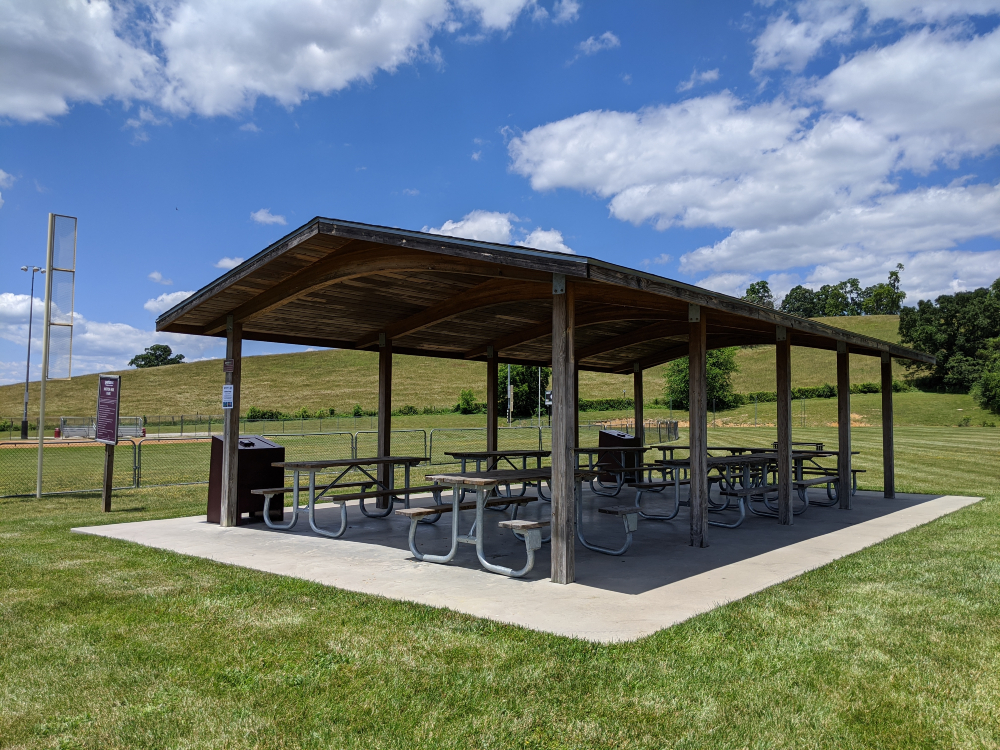 The picnic shelter at Motor Mile Park is shown located next to the baseball field and open green space on a sunny summer day.