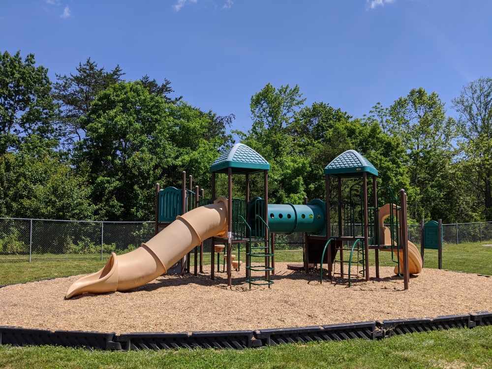 The playground at Plum Creek Park consists of slides and climbing areas on a mulch base bordered by green space and trees.