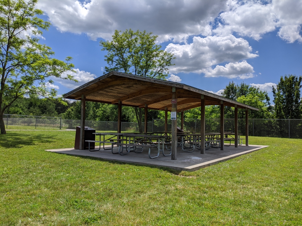 The picnic shelter at Plum Creek Park is shown in open green space with trees and the nearby baseball field on a sunny summer day.